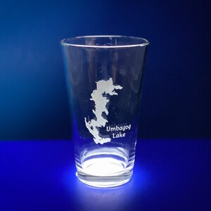 May include: A clear glass with an etched design of the outline of Umbagog Lake. The glass is on a blue surface.