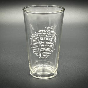 May include: A clear glass pint glass with a white design of a list of mountain names, including Marcy, Haystack, Whiteface, and Algonquin.