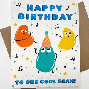 May include: A birthday card with the words "HAPPY BIRTHDAY" in blue. The card features cartoon beans in orange, yellow, and teal, with musical notes and confetti. The text "TO ONE COOL BEAN!" is at the bottom.