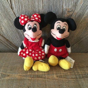 K&ouml;nnte beinhalten: Ein Paar Pl&uuml;schtiere von Mickey Mouse und Minnie Mouse. Mickey tr&auml;gt rote Shorts und schwarze Schuhe. Minnie tr&auml;gt ein rotes Kleid mit wei&szlig;en Punkten und gelbe Schuhe. Beide haben schwarze Augen und einen roten Mund.