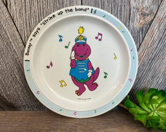Könnte beinhalten: Ein weißer Plastikteller mit blauem und grünem Rand, auf dem Barney der Dinosaurier als Bandleader dargestellt ist. Der Teller sagt "Barney TM says "Strike up the band"!"