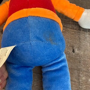 Puede incluir: Un juguete de peluche azul y naranja con una camisa roja. El juguete tiene una etiqueta de Disney pegada a su pierna.