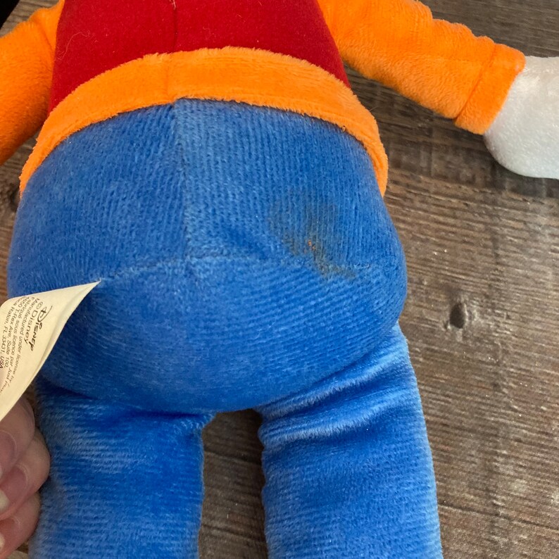 Puede incluir: Un juguete de peluche azul y naranja con una camisa roja. El juguete tiene una etiqueta de Disney pegada a su pierna.