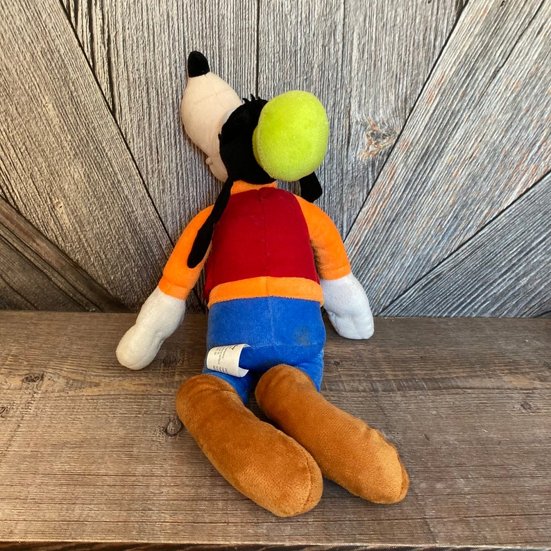 Puede incluir: Un juguete de peluche de Goofy, un personaje de dibujos animados de Disney. Lleva un chaleco rojo, una camisa naranja, pantalones azules y un sombrero verde. Est&aacute; tumbado de lado con las piernas dobladas.