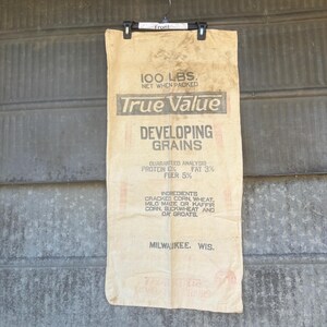 May include: A vintage True Value Developing Grains sack, cream-colored with black and red text. The bag reads "100 LBS. Net When Packed" and lists ingredients like cracked corn and wheat. The text also includes the city of Milwaukee, Wisconsin.
