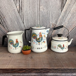 May include: A set of three white enamelware kitchen items with black handles and a rooster design. The set includes a creamer, a coffee pot, and a small kettle. The coffee pot has the words "Good Morning" and "Coffee Time" on it.