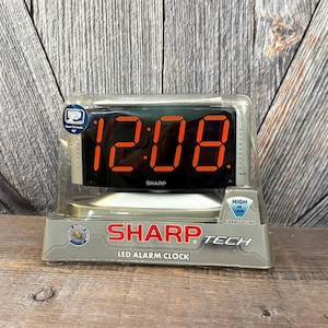 May include: A Sharp brand LED alarm clock in its original clear plastic packaging. The clock features a black display with bright red digital numbers showing 12:08. The packaging includes text labels for Sharp Tech, LED Alarm Clock, and High or Low alarm volume settings.