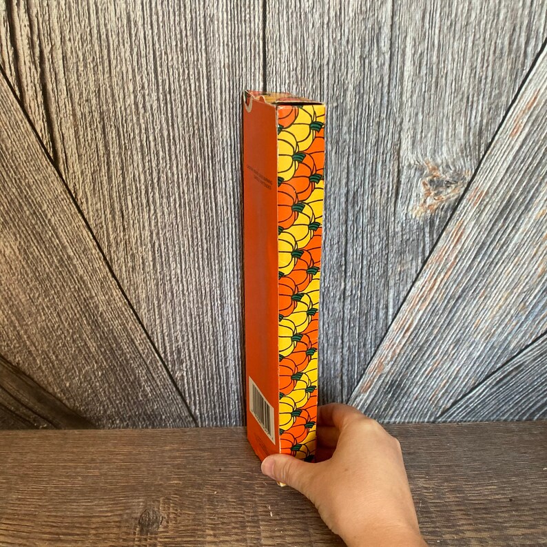 May include: An orange rectangular box with a repeating pattern of yellow and orange pumpkins. The box is held by a hand and is set against a wooden background.