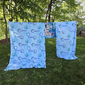 May include: Light blue Toy Story-themed bedding set hanging outside. The set features repeating patterns of Woody and Buzz Lightyear in blue circles. A matching blanket with the characters is also visible.