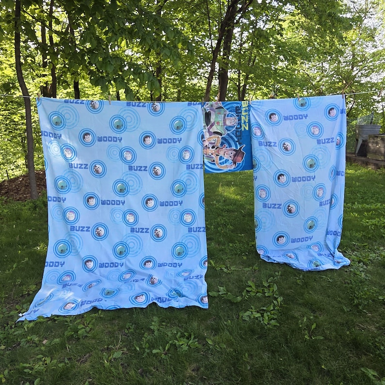 May include: Light blue Toy Story-themed bedding set hanging outside. The set features repeating patterns of Woody and Buzz Lightyear in blue circles. A matching blanket with the characters is also visible.