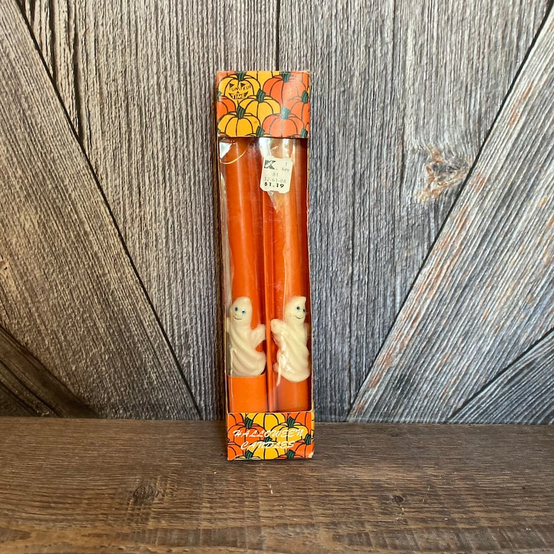 May include: A package of two orange taper candles with white ghost figures. The packaging features a pumpkin design and the words "Happy Halloween". The candles are displayed on a wooden surface.