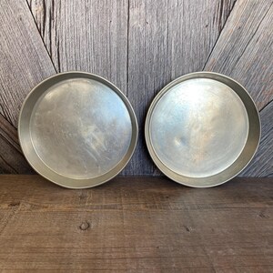 May include: Two round, silver-colored metal cake pans. The pans are empty and have a slightly worn appearance. They are sitting on a wooden surface with a wooden plank background.