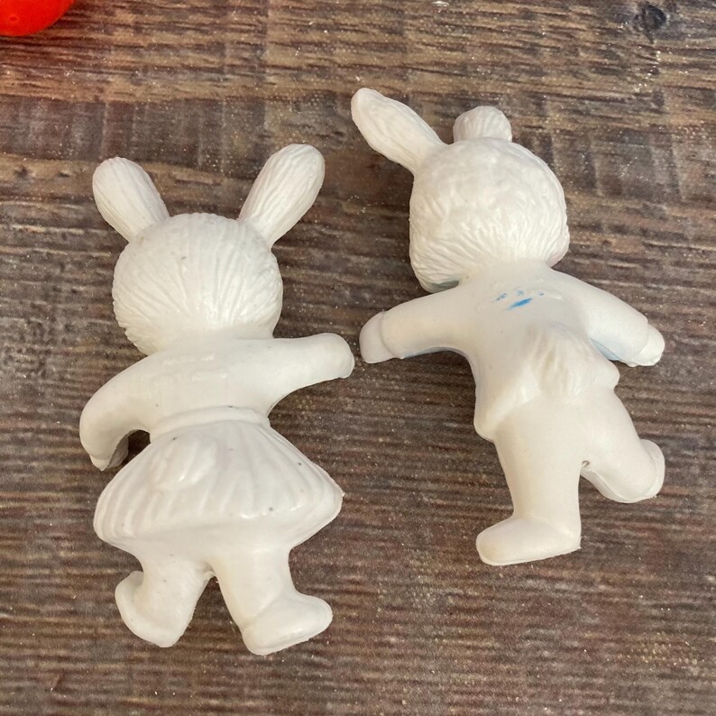 May include: Two white plastic bunny figurines. One bunny is wearing a dress and the other is wearing pants. The bunnies are facing opposite directions.