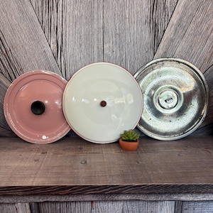 May include: Three vintage metal pot lids in a row. The lids are in different colors: pink, white, and silver. The pink lid has a black knob, the white lid has a red rim and a brown knob, and the silver lid has a handle.