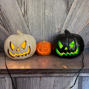 May include: Three illuminated jack-o'-lanterns in varying sizes. A large off-white pumpkin with yellow light, a small orange pumpkin, and a large black pumpkin with green light. All have carved faces and sit on a wooden shelf, perfect for Halloween.