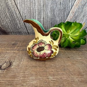 May include: A small, hand-painted ceramic pitcher with a cream-colored base. The pitcher features a large, stylized floral design in shades of burgundy and brown. The handle and spout are brown, with a green rim. A decorative piece.