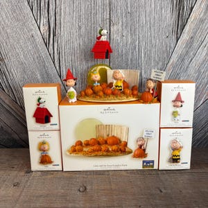 May include: A collection of Hallmark Keepsake ornaments featuring characters from the Peanuts comic strip. The ornaments include Snoopy's doghouse, Linus, Charlie Brown, and Sally, with a pumpkin patch theme. The boxes are orange and white.