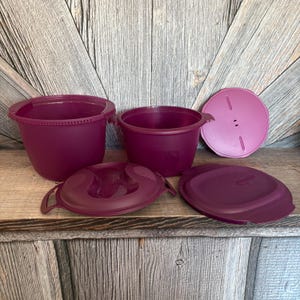 May include: A set of plum-colored plastic kitchen containers with lids. The set includes two round containers and two lids, one with a handle and one with vents. The containers are arranged on a wooden surface.