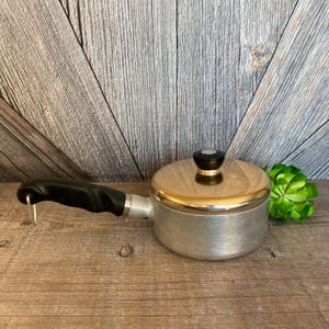 May include: A vintage aluminum saucepan with a copper lid and a black handle. The saucepan has a black knob on the lid.