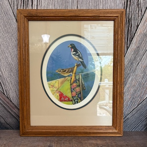 May include: A framed print featuring two birds perched on a decorative item. The artwork is set within an oval shape against a blue backdrop with trees. The frame is made of wood.