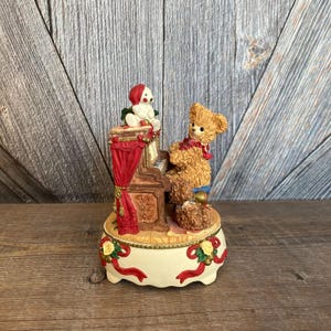 May include: A vintage-style musical figurine of a teddy bear playing a piano with a smaller teddy bear sitting on the piano bench. The figurine is decorated with red, green, and gold accents and has a white base.