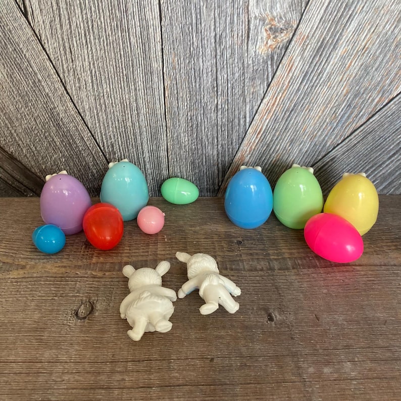 May include: A set of pastel plastic Easter eggs with bunny ears, including a pink, yellow, light blue, green, and purple egg. There are also two white plastic bunny figurines and a few smaller plastic eggs in red, blue, and green.