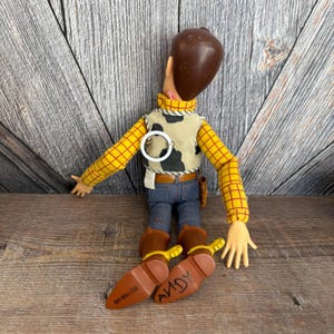May include: A Woody doll from the Toy Story franchise. The doll has a brown molded head, a yellow plaid shirt, a cow-print vest, blue jeans, and brown boots. The word "ANDY" is written on the bottom of the boots.