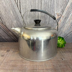 May include: A vintage stainless steel tea kettle with a black handle and lid knob. The kettle has a spout for pouring and a rounded body. The handle is arched and the lid sits on top. The kettle is sitting on a wooden surface.