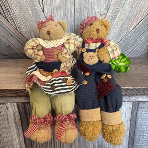 May include: A pair of plush teddy bears dressed in country-style clothing. The female bear is wearing a red and white plaid shirt, a blue and white striped skirt, and a red bow. The male bear is wearing a blue overall with a red and white plaid shirt and a red and white plaid hat.