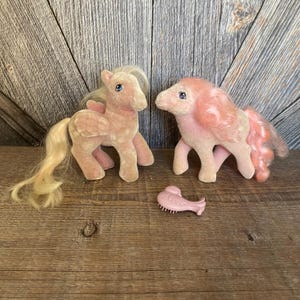 May include: Two pink plush My Little Pony toys with blonde and pink hair. The ponies are standing on a wooden surface. A pink plastic comb is in the foreground.