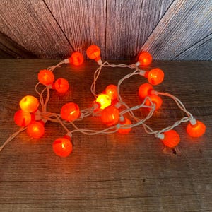 May include: A string of orange pumpkin-shaped lights with white wiring. The lights are strung together in a random pattern and are lit up.