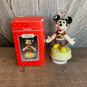 Puede incluir: Una figura de caja de m&uacute;sica vintage de Mickey Mouse de Disney en una caja roja. La figura est&aacute; hecha de porcelana y representa a Mickey Mouse con un disfraz marr&oacute;n y morado. La caja de m&uacute;sica reproduce una melod&iacute;a cuando se da cuerda.