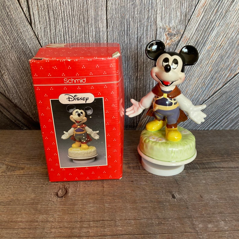 Puede incluir: Una figura de caja de m&uacute;sica vintage de Mickey Mouse de Disney en una caja roja. La figura est&aacute; hecha de porcelana y representa a Mickey Mouse con un disfraz marr&oacute;n y morado. La caja de m&uacute;sica reproduce una melod&iacute;a cuando se da cuerda.