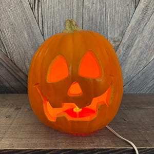 May include: An orange, illuminated jack-o'-lantern with a carved face. The pumpkin has triangular eyes, a triangular nose, and a wide, toothy grin. The interior glows with an orange light, and a white cord extends from the base.