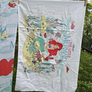 Może przedstawiać: Biała poszwa na pościel z kolorową ilustracją Arielki z Małej Syrenki Disneya. Projekt zawiera Arielkę, ryby, koralowce i tytuł filmu. Prześcieradło wisi na sznurku na zewnątrz.