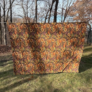 May include: A brown fabric with a repeating pattern of sunflowers, pumpkins, and grapes. The fabric is hanging on a clothesline outdoors, with a background of trees and grass. The colors are warm, autumnal tones.