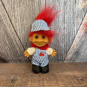 May include: A vintage troll doll with orange skin, red hair, and a blue and white striped hat. The doll wears a red scarf, a white shirt, and blue and white striped overalls with a red train graphic. Black shoes complete the outfit.