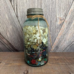 May include: A vintage blue glass jar filled with a variety of buttons in different colors and sizes. The jar has a metal lid and is tied with twine. The buttons range from white to black and are of various shapes.