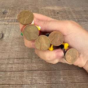 May include: A collection of vintage Lincoln pennies, each with a copper-brown hue, are arranged in a circular pattern. The pennies are attached to a colorful, cartoon-like figure with yellow, red, and black accents. The coins are held in a hand.