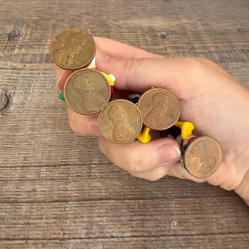 May include: A collection of vintage Lincoln pennies, each with a copper-brown hue, are arranged in a circular pattern. The pennies are attached to a colorful, cartoon-like figure with yellow, red, and black accents. The coins are held in a hand.