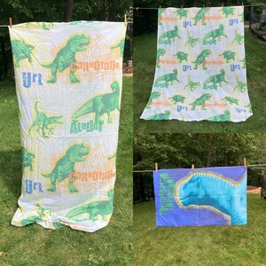 May include: Three dinosaur themed bed sheets. The first sheet is white with a green and orange dinosaur print. The second sheet is white with a green dinosaur print. The third sheet is blue with a blue dinosaur print.