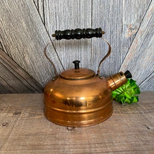 May include: A vintage copper tea kettle with a black wooden handle and a black plastic whistle. The kettle has a shiny copper finish and is sitting on a wooden surface.