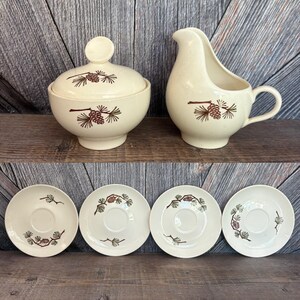May include: A vintage cream-colored ceramic sugar bowl with a lid, a creamer, and four saucers. Each piece is decorated with a brown pine cone and branch design. The set is displayed on a wooden surface.