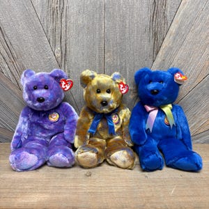May include: Three plush teddy bears in shades of purple, gold, and blue sit side-by-side. Each bear has a ribbon, a tag, and a circular emblem. The bears are on a wooden surface with a wooden plank background.