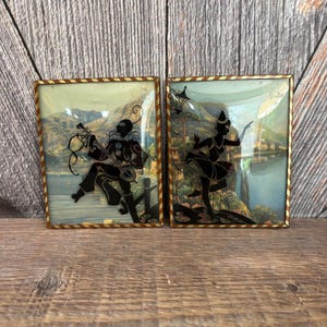 May include: Two framed vintage silhouette art pieces. One features a musician playing an instrument, the other a dancer. Both have black silhouettes against a scenic background with gold-toned frames. The frames have a brown and cream striped border.