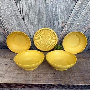 May include: A set of five vintage yellow plastic bowls and a matching lid. The bowls are round with a slightly flared rim. The lid is circular with a sunburst design. The bowls and lid are made of durable plastic.