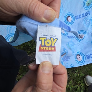 May include: A light blue fabric with a pattern featuring Woody and Buzz Lightyear from Toy Story. The tag shows the Disney Pixar Toy Story logo in yellow, red, and white.