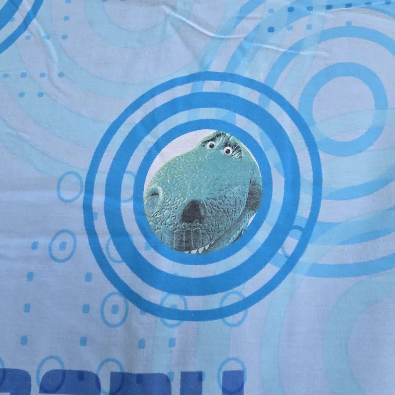 May include: A light blue bed sheet featuring a cartoon dinosaur character within a blue circular design. The background has a pattern of light blue concentric circles and dots. The dinosaur has a teal color with white eyes and a black nose.
