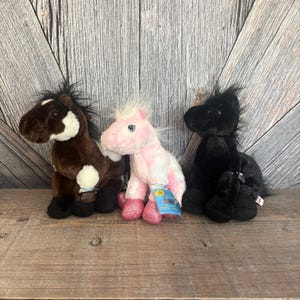 May include: Three plush toy horses in a row. The first is brown with a white patch and black mane. The second is pink with a white mane and pink hooves. The third is black with a black mane. All three are sitting on a wooden surface.