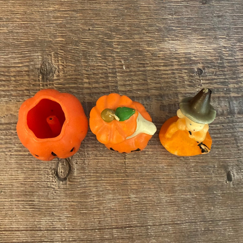 May include: Three orange pumpkin-shaped candles, one with a green leaf on top, one with a witch face and hat, and one with a hollow center.